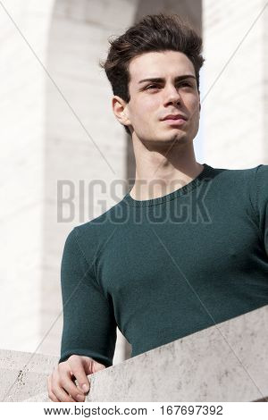 Stylish hair young man outdoors on the ledge, tight knit. A beautiful young European (Italian) man is outdoors resting on the ledge. The boy wears a tight knit long-sleeved green.