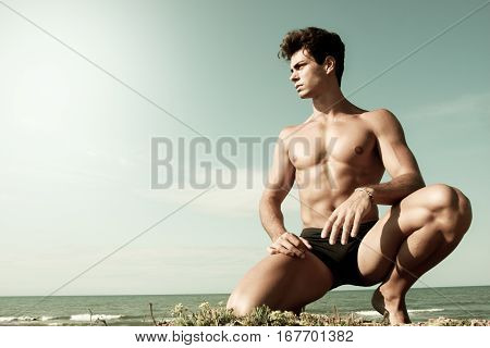 Young man naked on his knees. Sea and sky behind. A young man with a beautiful body is on his knees with the sea behind. Intense light and color antiqued.