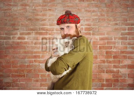 Young bearded man in plaid cap with fluffy cat on brick wall background