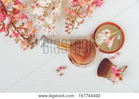 There White and Pink  Branches of Chestnut Tree,Brown Powder with Mirrow and Make Up Brush are on White Table,Top View