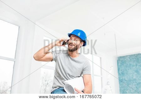 Handsome smiling repairman or builder in helmet talking with phone with drawings in the white interior