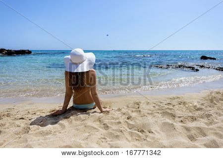 Young woman sitting on the beach. Portrait of a girl in white hat relaxing at tropicon beach. Beautiful Summer sea side beach with turquoise water. View from behind