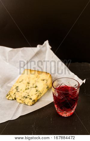 Shot glass with slow gin beside a wedge of stilton cheese unwrapped greaseproof paper.