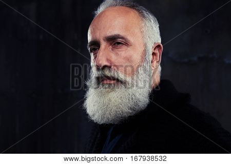 Close-up of a man wearing shirt and pullover, looking at camera against dark background. Man isolated in studio. Senior man with gray hair and beard