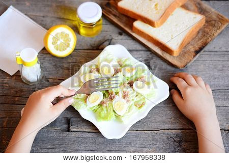 Small child holds a fork in his hand. Child eats salad with chinese cabbage, canned tuna and quail eggs. How to teach a kid to eat healthy food. Bread, lemon, olive oil jar on a vintage wooden table