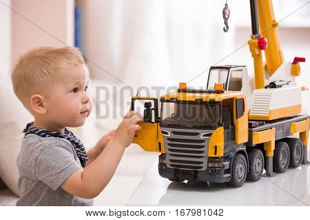 Portrait of adorable smiling toddler boy sitting at the table playing with a big construction car at home. Little child with toy car. Educative toys for children. Early learning and developing concept