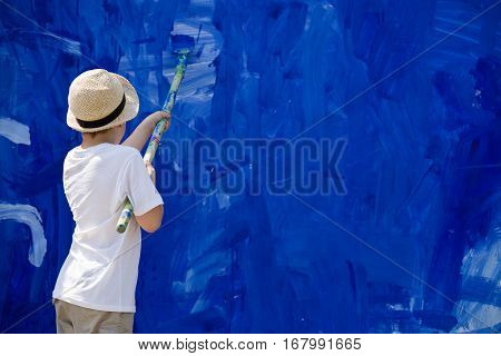 Cute kid boy in straw hat painting fence with blue color. Child drawing outdoors.