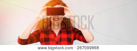 Young Red-haired Girl In A Red Checkered Shirt. A Young Girl Covered Her Eyes With A Phone