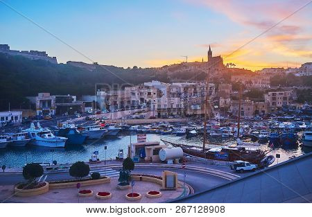 Ghajnsielem, Malta - June 15, 2018: The Scenic Fishing Harbor With Boats, Rocky Hills And Tall Silho