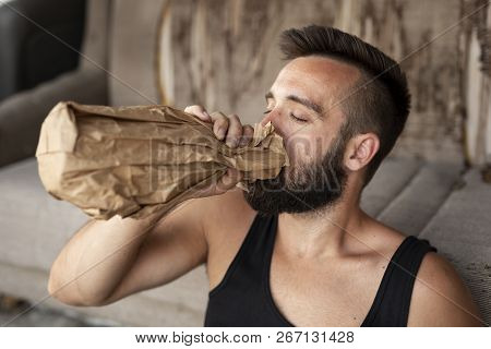 Drunkard Sitting On A Building Rooftop Terrace, Holding A Bottle In A Paper Bag, Drinking