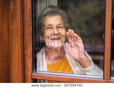 An Elderly Woman Standing By The Window, Looking Out. Shot Through Glass.