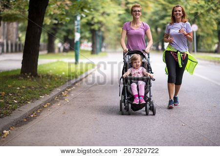 two young healthy women jogging together while pushing a baby stroller at city park
