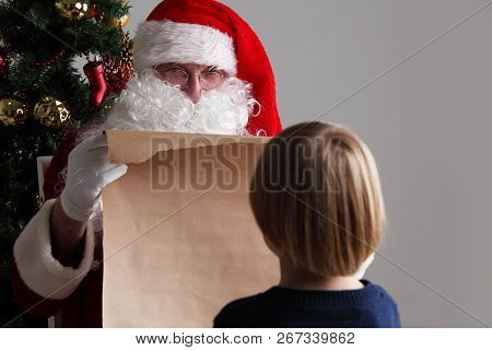 Santa Claus With With List Or Nice Or Naugthy List Paper With Small Boy Near Christmas Tree