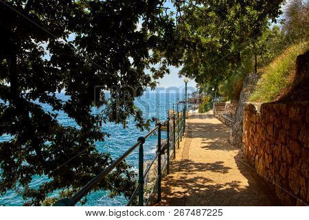 Lovran, Istria, Croatia. Picturesque embankment along resort old town at coast of Adriatic Sea with blue water and waves. Trees with green leaves. Warm sunny summer landscape.