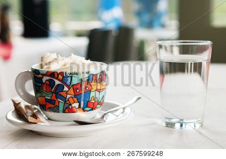 A Colorful Cup Of Coffee With Overflowing Whipped Cream, Sugar And A Glass Of Water On A Table With 