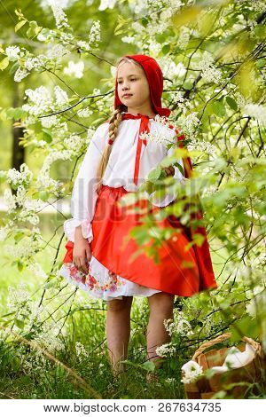Smiling Blond Girl Posing In A Dress Of Little Red Riding Hood Near Green Trees