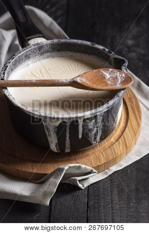 Boiled Milk In A Black Saucepan And Spilled Over The Edges, A Wooden Spoon, On A Cutting Board, On A