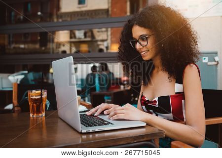 Cute Curly Hipster Female Freelancer In A Street Bar With Spritz Cocktail Working On Her Netbook; Dr