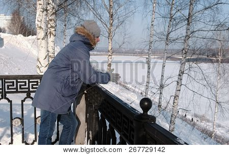 Man In Blue Down Jacket With Fur Hood Stands At The Fence And Look Around In The Winter Park.