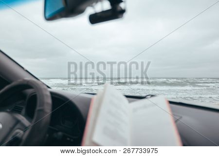 Book on car dashboard over sea waves outside. Weekend trip in bad rainy weather. Dramatic winter travel concept.