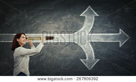 Positive, Young Woman Looking Through A Spyglass In Front Of A Blackboard As A Crossroad Arrows Spli