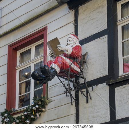 A Model Of Father Christmas Sitting On A Chair Reading A Book, Which Is Hanging On A Building Wall