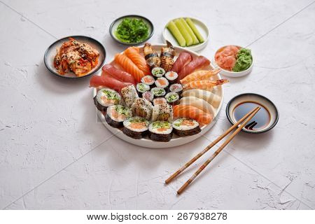Asian food assortment. Various sushi rolls placed on round ceramic plate. Kimchi and goma wakame salads, fresh cucomber, ginger and wasabi. Soy souce and chopsticks on sides.