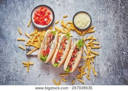 Assortment of three tasety hot dogs, placed on wooden cutting board. Served with french fries, onion and tomato. View from above. Stone background.