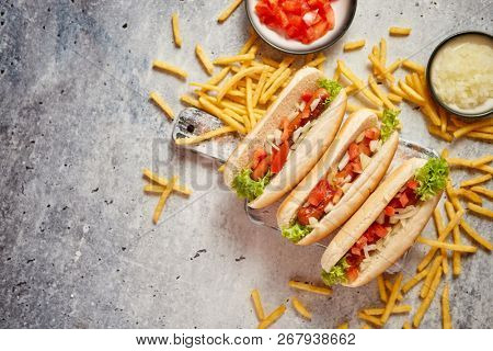Assortment of three tasety hot dogs, placed on wooden cutting board. Served with french fries, onion and tomato. View from above. Stone background.