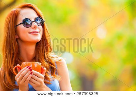 Style Redhead Girl In Sunglasss With Cup In The Autumn Park.