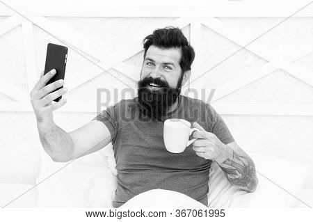 Source Of Energy. Bearded Man Enjoy Video Conferencing And Coffee In Bed. Happy Hipster Hold Cup And