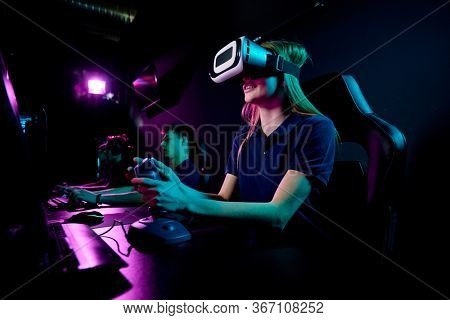 Contemporary eenage girl in vr headset using joystick while sitting in front of computer monitor and playing new video game in club