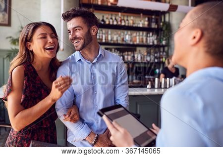 Couple Being Greeted By Maitre D Using Digital Tablet As They Arrive At Restaurant