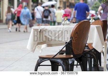 A Table With A White Tablecloth Small Red Flowers On It, Wooden Chair In Front, People Walking On Th
