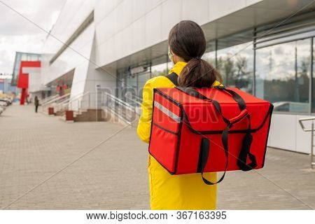 Rear view of young female courier carrying big red bag with orders of clients while walking along modern supermarker exterior outdoors