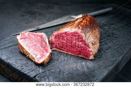 Barbecue dry aged beef fillet head medallion steak natural with a knife as closeup on a charred wooden board 
