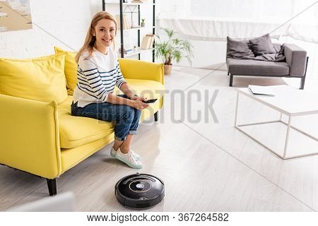 Beautiful Woman With Smartphone Smiling And Looking At Camera On Sofa Near Coffee Table And Robotic 
