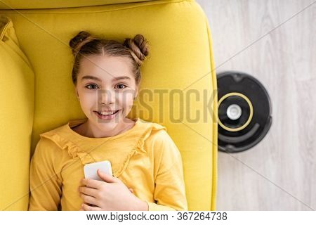 Top View Of Cute Kid With Smartphone Smiling, Looking At Camera And Lying On Sofa Near Robotic Vacuu