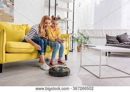 Happy Woman And Kid With Smartphone Smiling And Looking At Robotic Vacuum Cleaner Near Coffee Table 