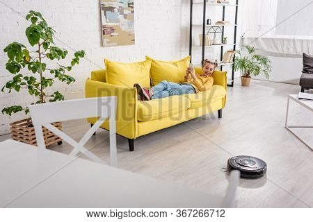 Selective Focus Of Kid On Sofa Looking At Robotic Vacuum Cleaner On Floor In Living Room
