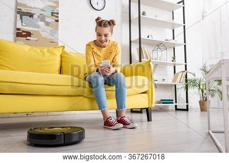 Cute Child With Smartphone On Sofa Smiling And Looking At Robotic Vacuum Cleaner On Floor In Living 