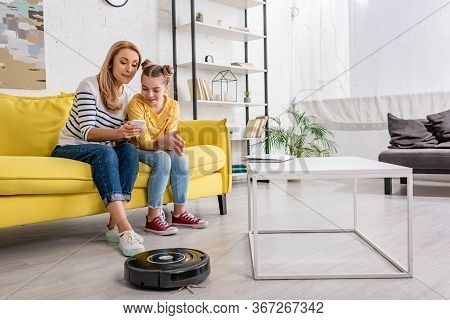 Mother And Daughter Looking At Smartphone On Sofa Near Coffee Table With Laptop And Robotic Vacuum C