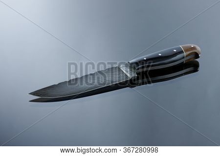 An All-metal, Middle Kitchen Knife Lies On A Mirror Surface, A Blade Made Of Multilayer Damask Steel