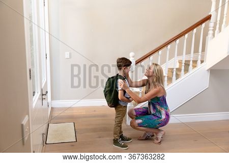Side view of a Caucasian woman kneeling in the hallway smiling and preparing her young son to leave for school, the son is wearing a backpack