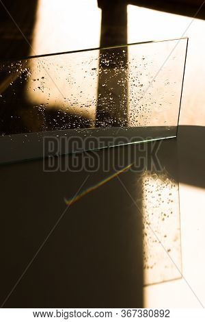 A Piece Of Glass With Water Drops Standing In Intence Sunlight And Reflecting A Rainbow On White Sur