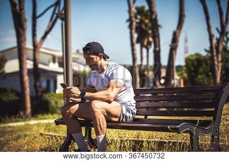 Sporty Man Personal Trainer Sitting Looking At His Phone On A Bench In An Outdoor Park. Muscle Body 