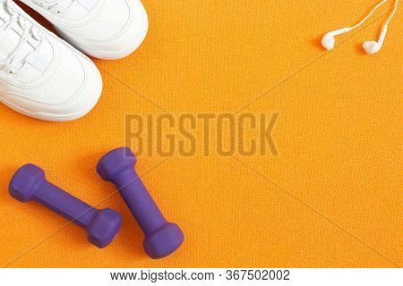 Sneakers, Dumbbells, Mobile Phone With Headphones On Background Of Orange Fitness Mat. Flat Lay Comp