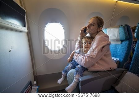 Young Beautiful Mother Is Sitting In An Airplane Chair And Talking On The Phone, And Hugs Her Little