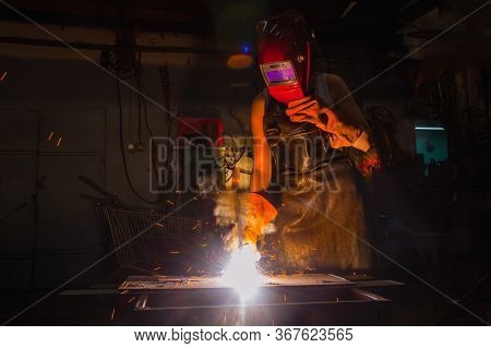 Female Welder Working In A Harsh Dark Workshop, Works With Steel With Sparks Flying.
