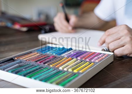 Person, Artist Painting With A Pastel Crayon Chalk On A White Sheet Of Paper And Pellette On The Woo